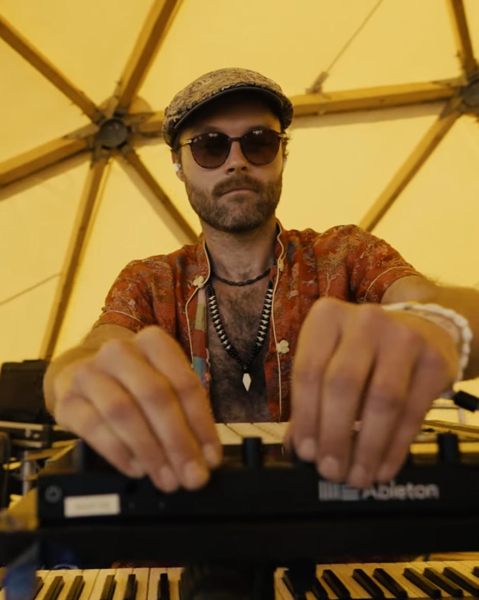 Keyboard player in a flat cap behind the Ableton rig in warm daylight