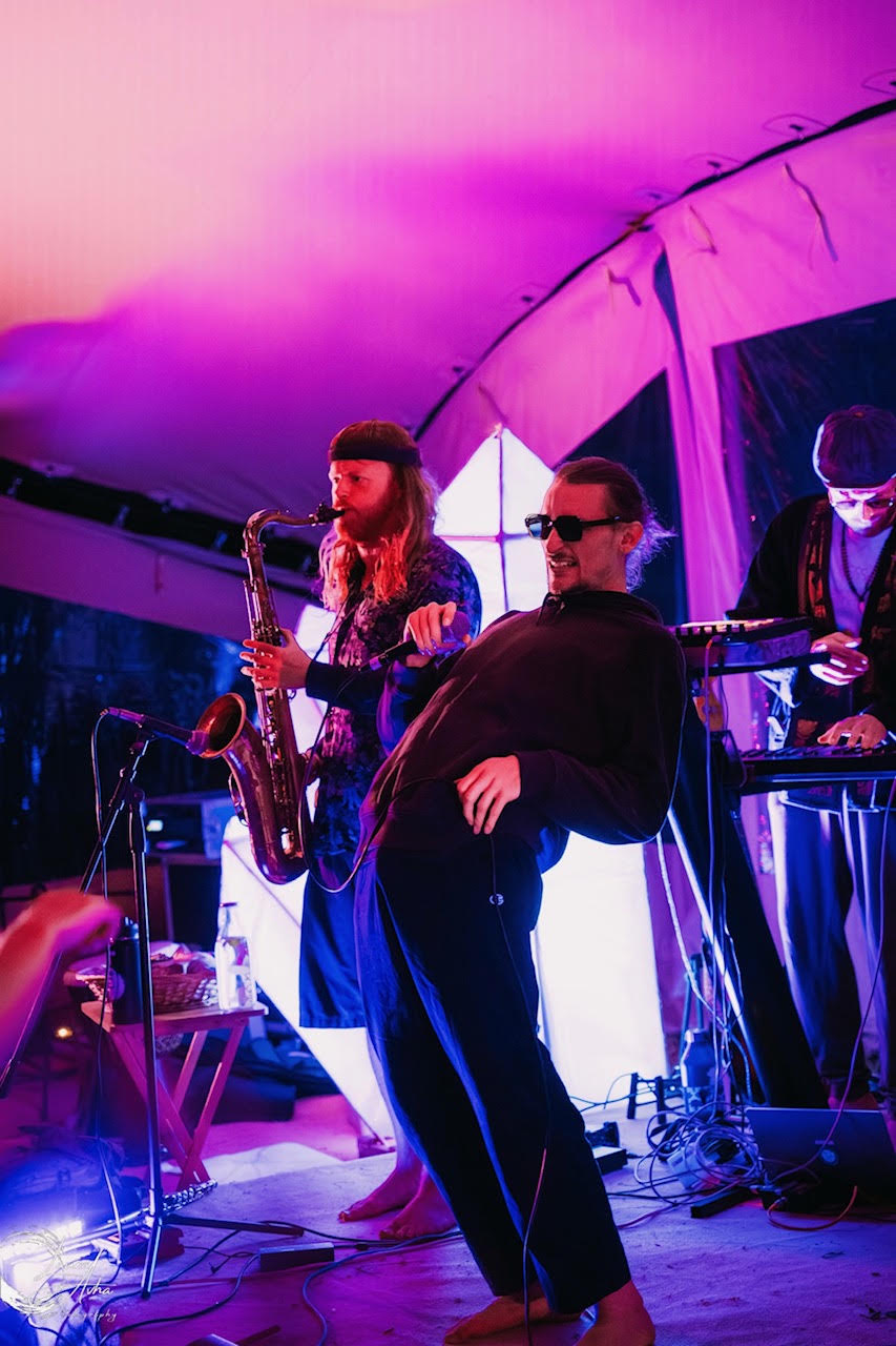 MC dancing barefoot in sunglasses beside saxophone player under pink light